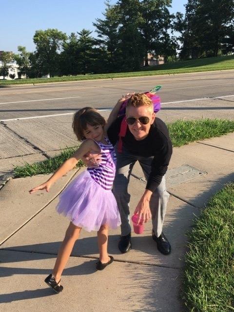 A parent clowning around on the sidewalk with a young girl in a purple tutu